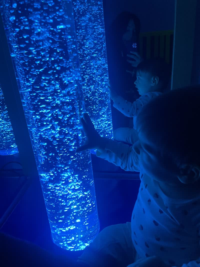 Un enfant touche une colonne à bulles lumineuse bleue dans la pénombre de la salle Snoezelen